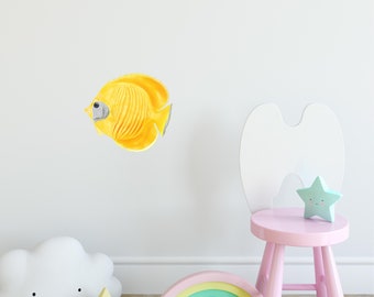 Op de afbeelding: Een kinderkamerscène met een gele vis muursticker, een witte wolkenlamp, een ster-vormig nachtlampje, een regenboog, een roze kruk en een sterdecoratie. De decoratie staat tegen een witte muur.