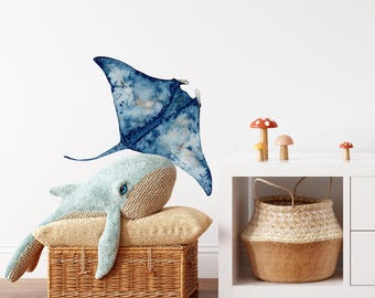 May include: A plush light blue whale toy rests on a tan cushion atop a woven wicker basket. Above the whale, a watercolor-style wall decal depicts a dark blue manta ray. Nearby, three small mushroom figurines sit on a white shelf with a basket.