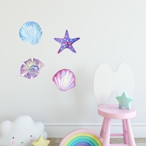 May include: A set of five watercolor-style wall decals featuring a starfish, seashells, and a flower. The decals are in shades of blue, purple, pink, and yellow. The decals are shown on a white wall with a pink chair and a rainbow.