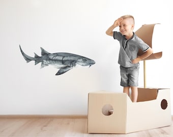 以下が含まれることがあります： グレーのシャツと青いショートパンツを着た子供が、ボートの形をした段ボール箱の中に立っています。子供は左を見て、片手を額に当てて、まるで海を見ているかのようです。子供の後ろの壁には、水彩画のグレーのシャークが描かれています。