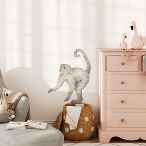 May include: A watercolor-style monkey wall decal in shades of gray and white, positioned above a woven basket with white polka dots. The scene includes a pink dresser, a gray armchair, and decorative flamingo figurines.