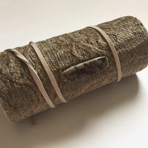 May include: A rolled-up, textured, olive-green fabric case secured with a light tan leather strap. A decorative stone clasp is centered on the case. The case is designed to hold items.