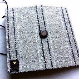 May include: A square, light gray fabric pouch with vertical navy blue stripes and a brown leather strap. The pouch has a dark brown button closure and a small, square leather accent with a gold stud.