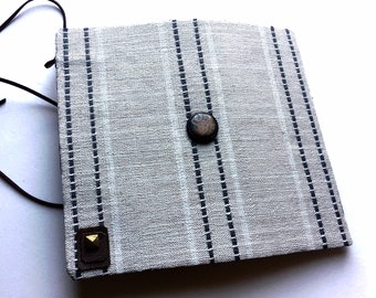 May include: A square, light gray fabric pouch with vertical navy blue stripes and a brown leather strap. The pouch has a dark brown button closure and a small, square leather accent with a gold stud.