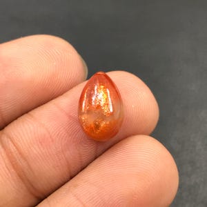 Puede incluir: Un cabujón de piedra de sol en forma de lágrima, de color rojo-naranja y con un acabado brillante e iridiscente.