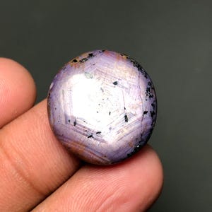 May include: Oval-shaped, purplish-pink gemstone with a star-like pattern. The gem has a polished surface with subtle color variations and small dark inclusions. The stone is held in a hand against a dark background.