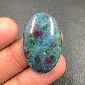 以下が含まれることがあります： 磨かれた表面を持つ楕円形の宝石。石は、ティール、緑、濃い赤など、さまざまな色を示しています。赤は石全体にパッチ状に現れ、ティールと緑はベースカラーを形成しています。