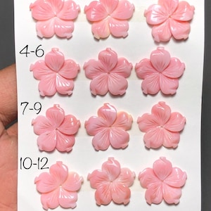 Puede incluir: Doce cuentas rosas en forma de flor, cada una con cinco pétalos y un diseño floral tallado, están dispuestas sobre una superficie blanca. Las cuentas están numeradas del 1 al 12. También se ve el texto "20m" y "12Pcs".