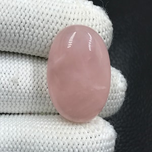 Pode incluir: Um cabochão de quartzo rosa oval, liso e de cor rosa claro. A pedra é polida e tem uma aparência translúcida.