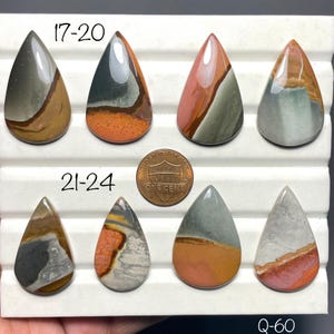 May include: Eight polished, teardrop-shaped jasper cabochons arranged in two rows, labelled 17-20 and 21-24. The stones feature varied patterns of grey, orange, brown, and cream. A US one-cent coin is placed in the centre for scale reference.