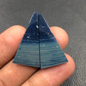 Puede incluir: Dos piedras preciosas triangulares de color azul oscuro con un efecto degradado, que transita de un azul profundo en la parte superior a un azul más claro en la parte inferior. Pequeñas motas blancas están dispersas por la superficie, pareciendo estrellas. Las piedras se sostienen en una mano.