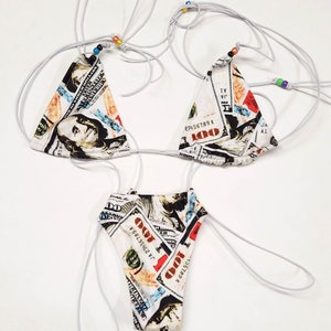 May include: A three-piece bikini set featuring a print of U.S. one hundred dollar bills. The triangle top and bottom are connected by thin, white cords. Small, colorful beads adorn the cords. The bikini is designed for swimwear.