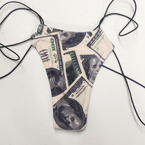 Puede incluir: Braga de bikini de cuerda negra con un estampado de billetes de dólar estadounidense. El estampado presenta el retrato de Benjamin Franklin y el texto "100" y "ONE HUNDRED DOLLARS".