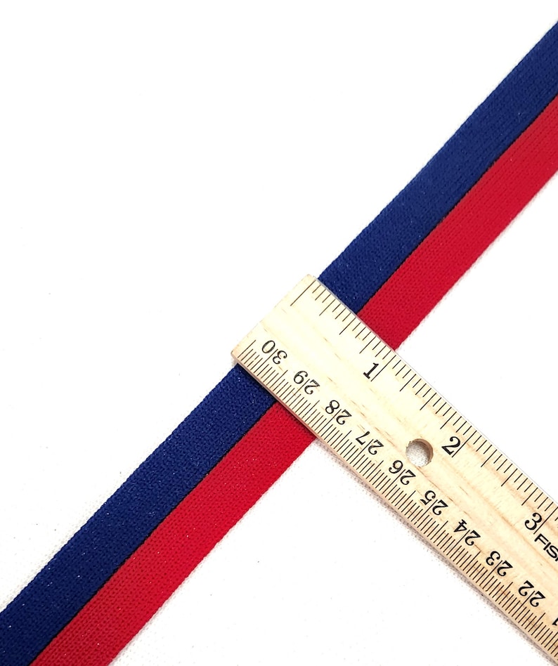 May include: Red and navy blue striped ribbon measured with a ruler. The ribbon is 1 inch wide.