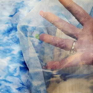 May include: A sheer white tulle fabric with a fine mesh pattern. The fabric is draped over a blue and white tie-dye fabric.