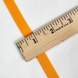 May include: A close-up of a bright orange elastic band measured with a wooden ruler marked in inches. The ruler reads "Made in U.S.A."