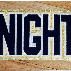 Könnte beinhalten: Ein rechteckiger Aufnäher mit dem Wort "KNIGHTS" in marineblauen Blockbuchstaben, weißen und goldenen Details. Der Aufnäher hat einen goldenen Glitzerrahmen, geeignet für ein Sportteam oder einen Verein.