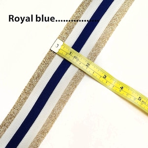 May include: A close-up of white fabric with gold and royal blue stripes. A yellow measuring tape is placed on top of the fabric, showing a measurement of 2 inches.