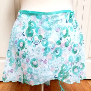 Puede incluir: Una falda de danza de gasa azul claro con un estampado de acuarela verde y morado de círculos y remolinos. La falda tiene una cintura elástica verde y un lazo en la parte posterior. El texto "Sweeping away by the beauty of it all" está impreso en la falda.