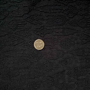 May include: Black fabric with a textured pattern of small, repeating designs. A gold coin is placed on the fabric.
