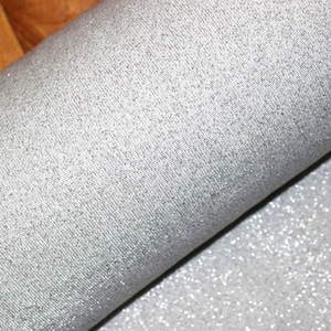 Może przedstawiać: Rulon srebrnej tkaniny brokatowej. Tkanina ma drobną, równomierną strukturę brokatu.