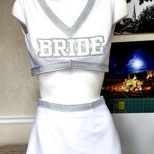 以下が含まれることがあります： 銀色のグリッターのトリムとトップに銀色のグリッターの文字で「BRIDE」と書かれた白いチアリーディングユニフォームです。