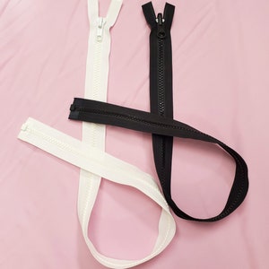 Può includere: Due cerniere lampo, una nera e una bianca, sono disposte su una superficie rosa. Entrambe le cerniere sono chiuse e hanno un cursore in metallo.