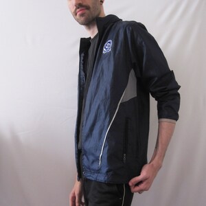 May include: A navy blue windbreaker jacket with a white zipper and white accents. The jacket has a logo on the left chest that says "C.U.N.Y.". The jacket is worn over a black t-shirt and black pants with a white Adidas logo.