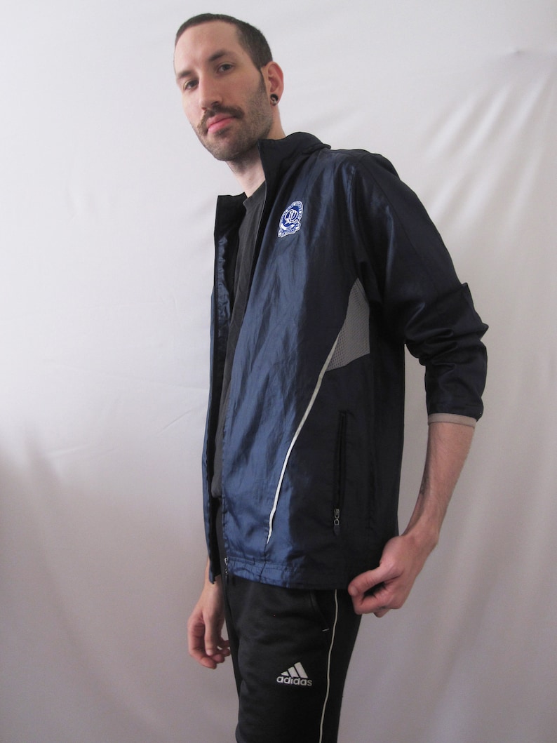 May include: A navy blue windbreaker jacket with a white zipper and white accents. The jacket has a logo on the left chest that says "C.U.N.Y.". The jacket is worn over a black t-shirt and black pants with a white Adidas logo.