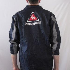 May include: A navy blue jacket with a gray and white stripe on the sleeves. The jacket has a red rooster logo in a white triangle with the text "le coq sportif" underneath.