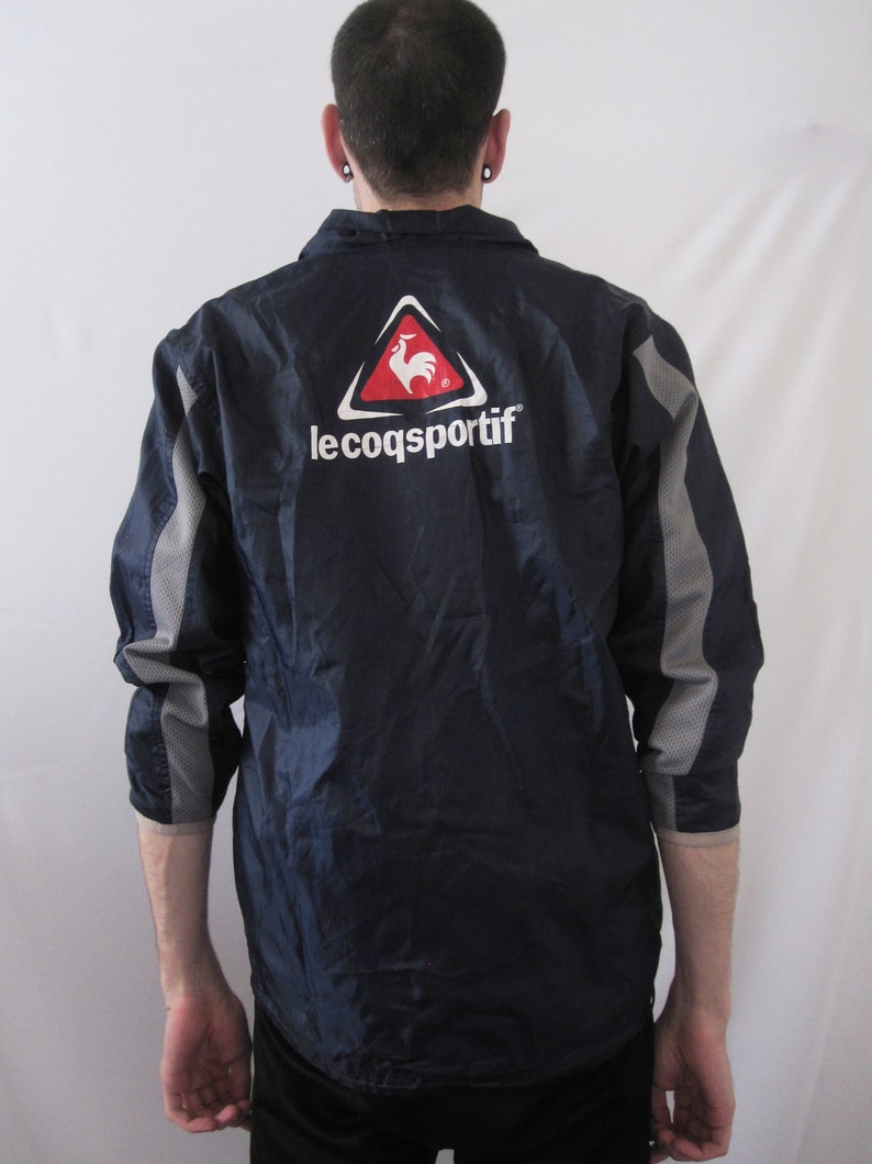 May include: A navy blue jacket with a gray and white stripe on the sleeves. The jacket has a red rooster logo in a white triangle with the text "le coq sportif" underneath.