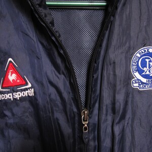 May include: A navy blue windbreaker jacket with a zipper closure. The jacket features a red rooster logo on the left side with the text "le coq sportif" and a blue and white logo on the right side with the text "Queens Park Rangers FC 1882 Loftus Road".