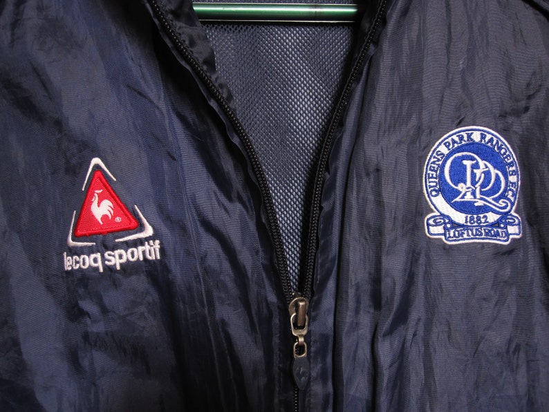 May include: A navy blue windbreaker jacket with a zipper closure. The jacket features a red rooster logo on the left side with the text "le coq sportif" and a blue and white logo on the right side with the text "Queens Park Rangers FC 1882 Loftus Road".