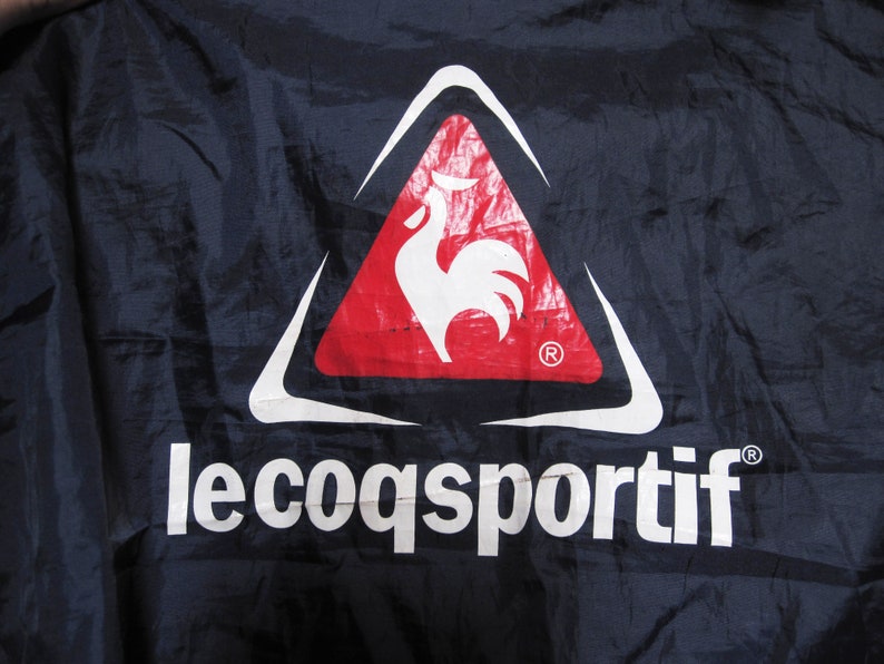 May include: A white rooster logo inside a red triangle with a white outline. The text "le coq sportif" is below the logo in white letters on a dark blue background.