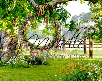 May include: A floral wreath hangs from a tree branch in a green grassy field. The wreath is made of greenery and purple and pink flowers. The text "By Michelle Photography" is visible in the background.