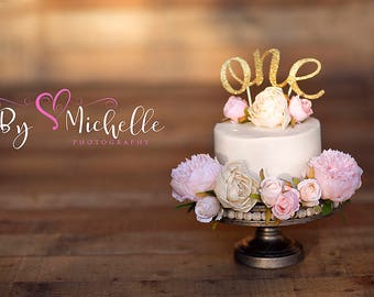 May include: A white cake with a gold glitter "one" topper and pink and white flowers on a cake stand. The cake is on a wooden surface with a watermark that says "By Michelle Photography".