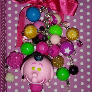 May include: A pink character keychain with a variety of colorful beads and a pink ribbon. The keychain features a smiling pink character with a small hat, surrounded by green, pink, and blue beads. The background is a purple box with white polka dots.