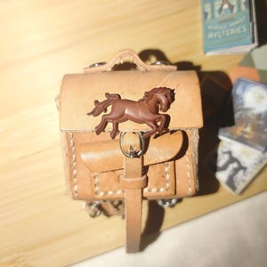 Puede incluir: Una pequeña mochila de cuero marrón con una hebilla en forma de caballo. La mochila tiene una correa y una solapa con una hebilla.