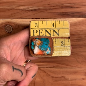 May include: A small, square piece of turquoise, orange, and white stone set in a brown leather frame. The frame is held within a yellow measuring tape marked with the word "PENN" and inch measurements.