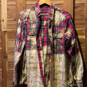 May include: A distressed plaid shirt in red, blue, and yellow, hanging on a wooden hanger. The shirt has a bleached, faded effect, with two chest pockets and a button-down front. The background is a wooden wall.