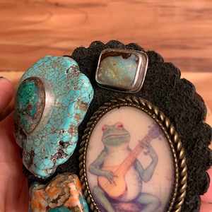May include: A collection of unique jewelry components, including a turquoise stone, a rectangular opal in a silver setting, and a small oval frame with a frog playing a ukulele. The components are set on a black, scalloped leather backing.