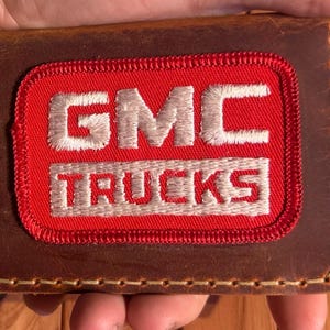 May include: A brown leather wallet with a red patch featuring the embroidered text "GMC TRUCKS" in white. The wallet has a stitched border and a distressed finish, showcasing a vintage aesthetic.