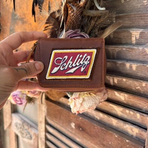 May include: A brown leather zippered coin purse featuring a rectangular patch with a red background and a white border. The patch displays the word Schlitz in a white cursive script font with a black outline and a small registered trademark symbol.