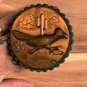 May include: A copper-colored round coaster with a desert scene featuring a roadrunner, cactus, and clouds. The coaster has a scalloped dark green border. The roadrunner is in motion, and the cactus is in the center.