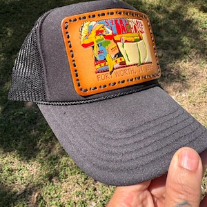 May include: A black trucker hat featuring a rectangular tan leather patch stitched with black thread. The patch displays a colorful graphic of a longhorn steer and the text 53RD NATIONAL LONGHORN SHOW FORT WORTH, TX 1993 in black lettering.