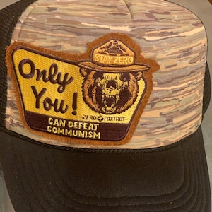 May include: A brown and black camouflage trucker hat with a brown patch featuring a cartoon bear wearing a ranger hat. The patch says "Stay Zero" and "Only You! Can Defeat Communism".