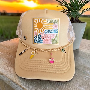May include: Tan trucker hat with a mesh back and a patch that reads "Forever Chasing Sunsets." The patch features a sun, flowers, and text. A gold chain with charms of a wine bottle, cocktail, guitar, and palm tree hangs from the brim.