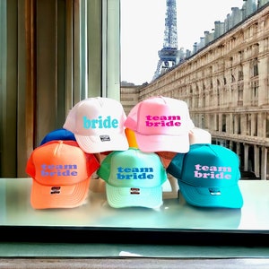 May include: Five trucker hats with mesh back panels. The hats are in various colors: pink, orange, blue, green, and white. The hats have the text "team bride" or "bride" in pink and blue lettering.