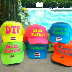 Puede incluir: Seis gorras de camionero de color ne&oacute;n con diferentes frases. Las gorras est&aacute;n dispuestas en semic&iacute;rculo sobre una superficie de madera. Las frases en las gorras son "DTF", "Shit Talker", "Bad & Boujee", "Dirty Dancer", "Rackout" y "Party Trick".