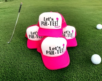 May include: A stack of four pink and white trucker hats with the text "Let's PAR-TEE!" and a golf tee graphic. The hats are on a green artificial grass surface with a golf club and two golf balls.